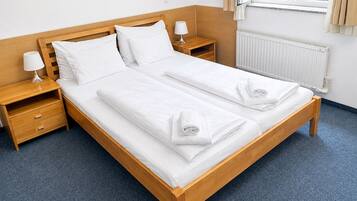 A double bed with white linens, a wooden headboard, two bedside tables with lamps, a radiator, and a window with curtains.
