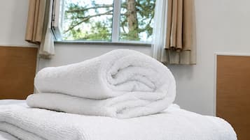 A neatly made bed with a stack of white towels, a window with curtains, and a view of trees outside.