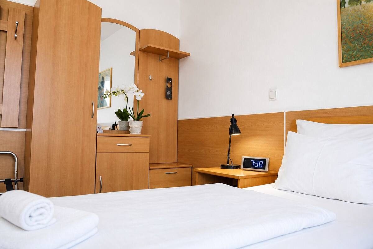 A hotel room with a bed, a nightstand with a clock, a dresser with a mirror, and a potted plant.