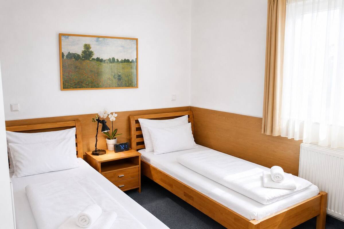 A hotel room with two single beds, a nightstand with a clock and a plant, a painting on the wall, and a window with curtains.
