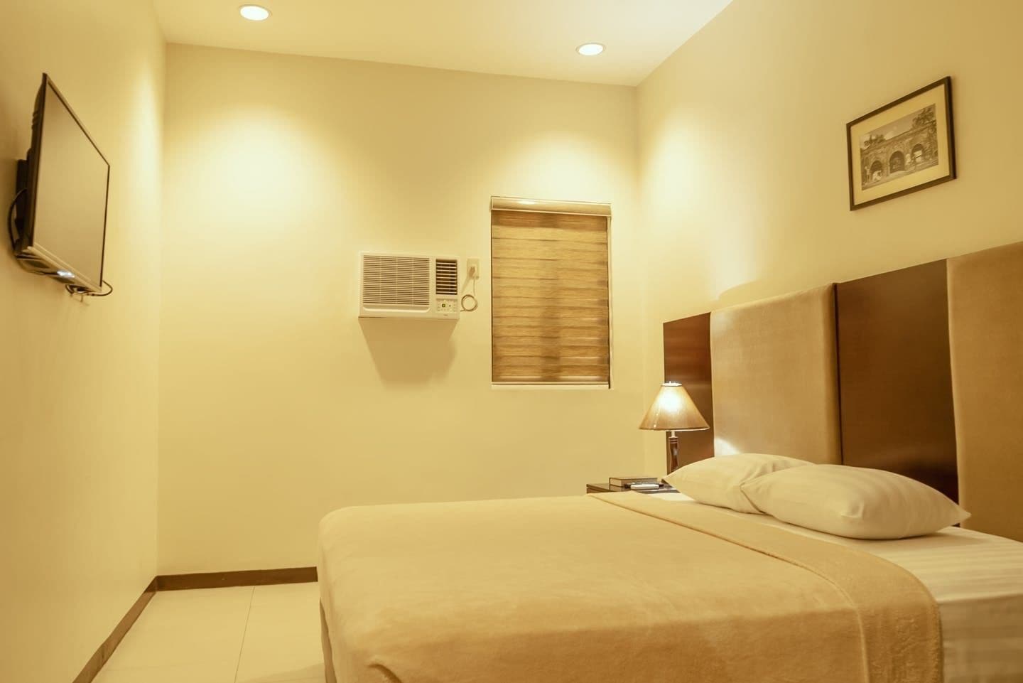 A hotel room with a bed, a bedside lamp, an air conditioning unit, a wall-mounted television, and a framed picture.