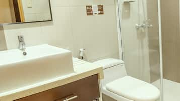 Un baño con un lavamanos blanco, un inodoro y una ducha con mampara de vidrio. Hay dos letreros en la pared, uno indica que no se permite fumar y el otro, que no se permite vapear.