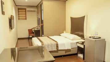 Ein Hotelzimmer mit Bett, Schreibtisch, Stuhl, kleinem Kühlschrank und einem Fenster mit Jalousien.