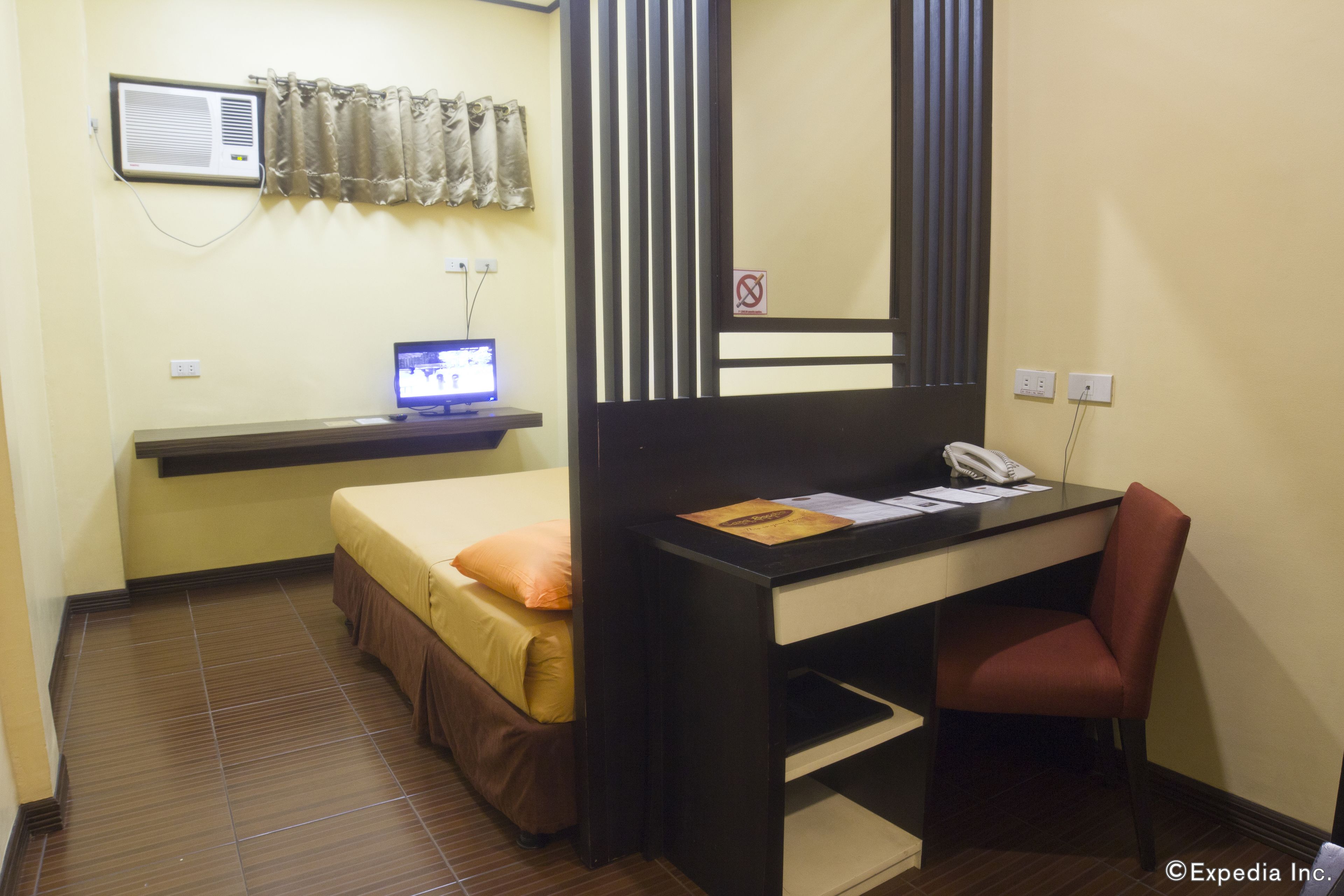 Habitación de hotel compacta con una cama individual, un escritorio con teléfono y un monitor de computadora.