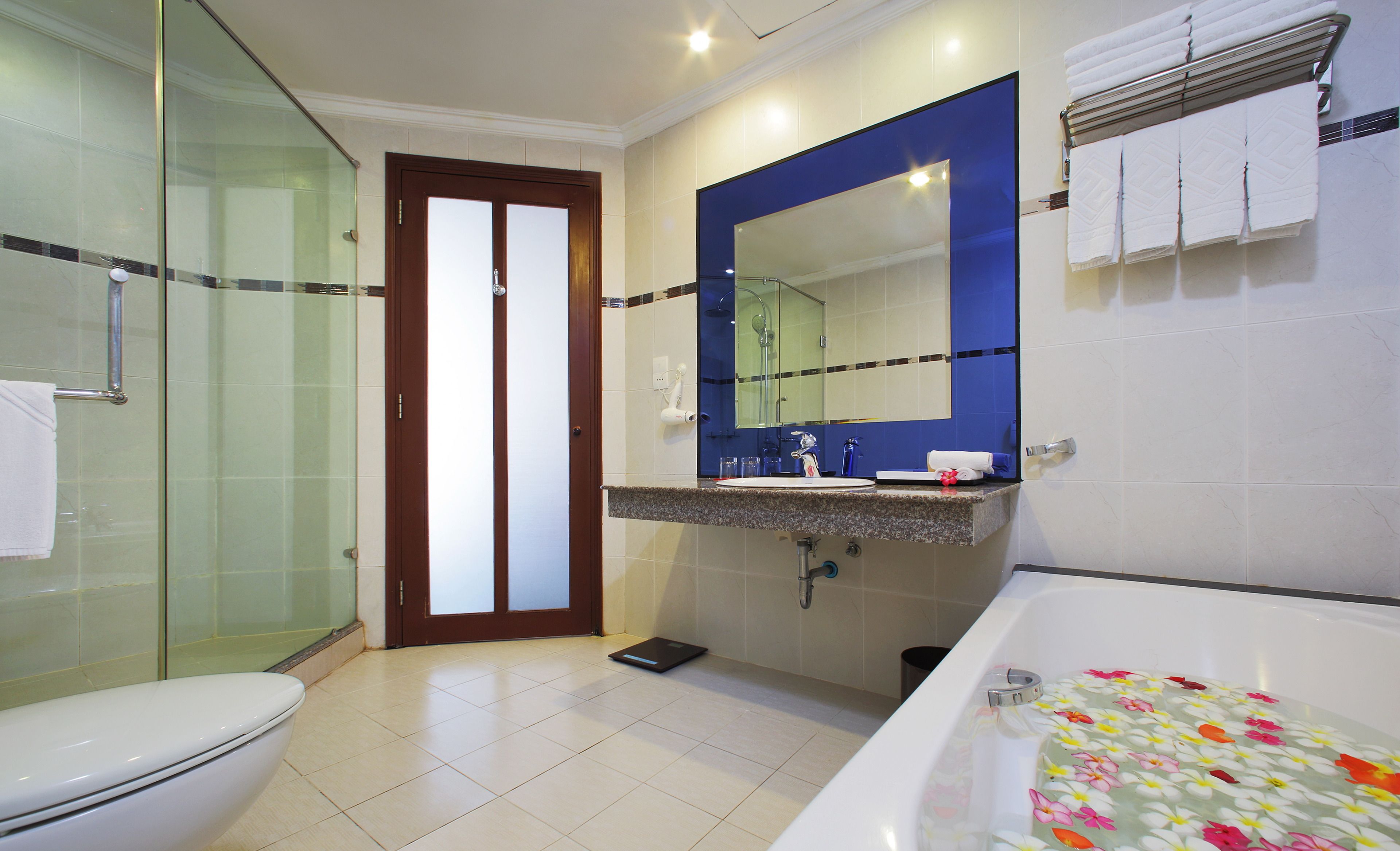 Un baño con cabina de ducha de vidrio, inodoro, lavabo con espejo y bañera con felpudos estampados de flores.