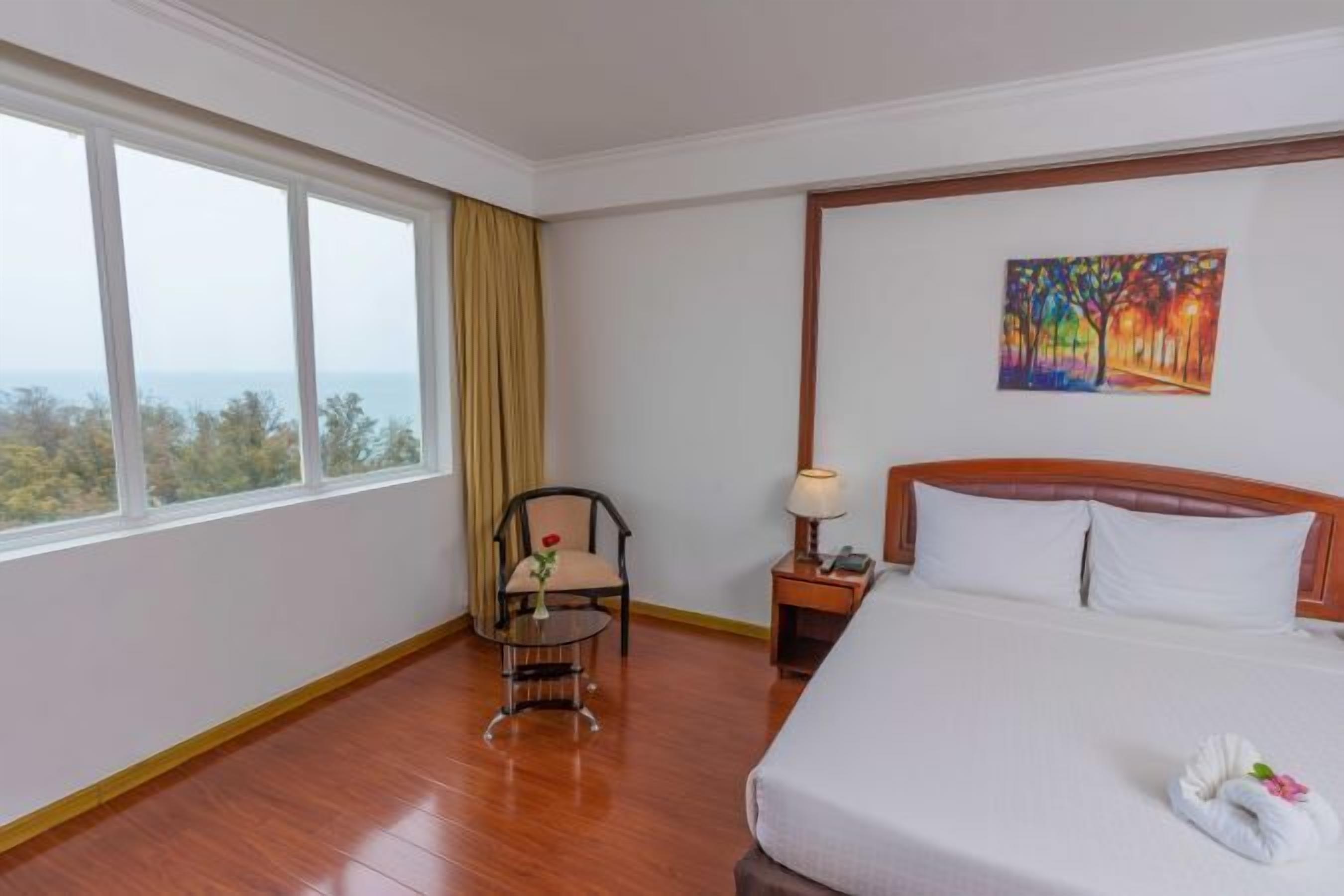 Una habitación de hotel con un ventanal grande, una cama con sábanas blancas, un cabecero de madera, una mesita con una silla y un cuadro en la pared.