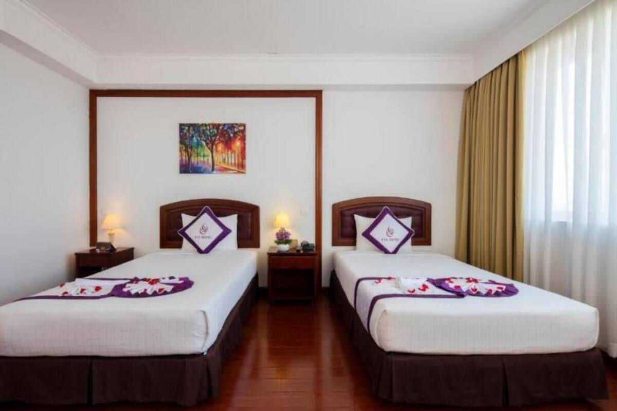 Une chambre d’hôtel avec deux lits simples, une table de chevet avec une lampe, un tableau au mur et une fenêtre avec des rideaux.