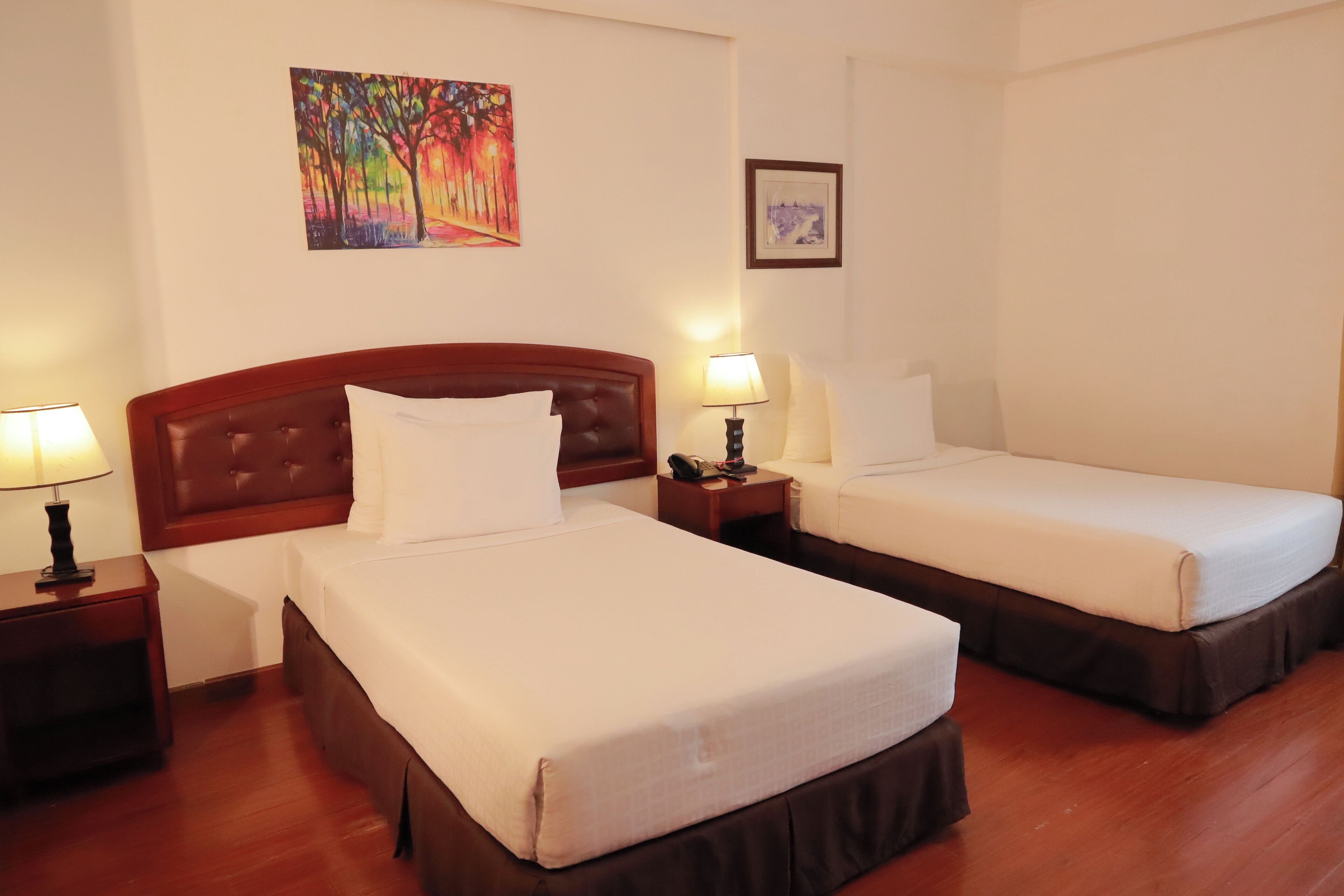 A hotel room with two beds, wooden headboards, white linens, a nightstand with a lamp, and a painting on the wall.
