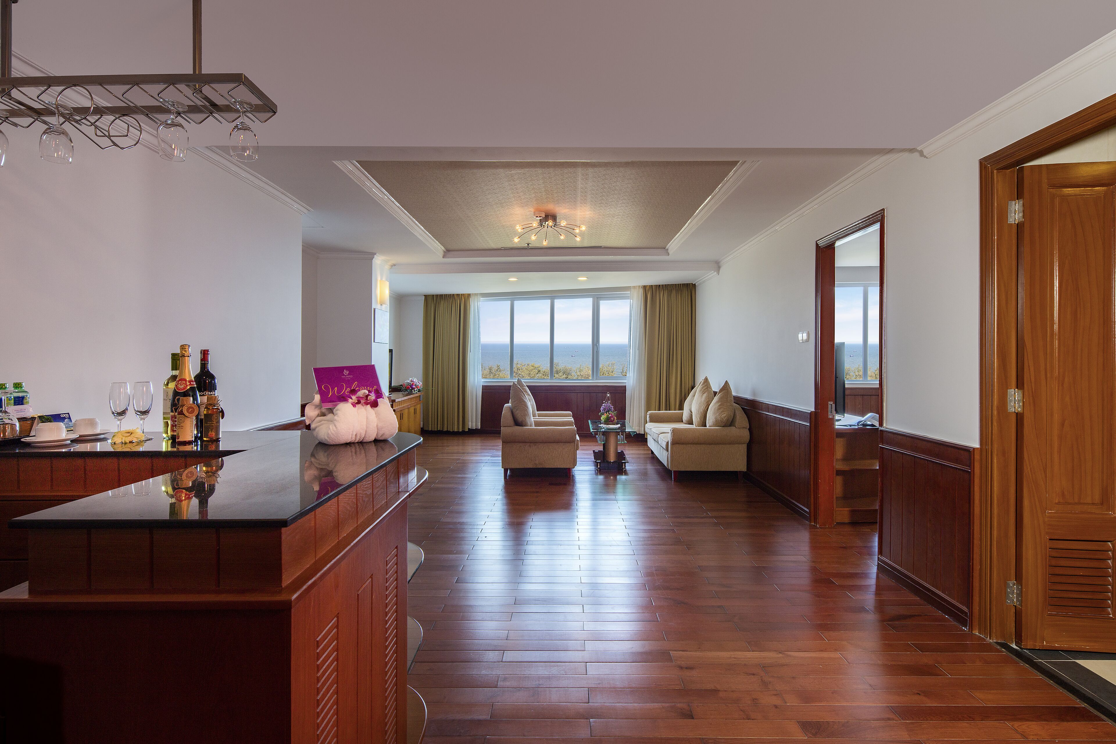 Un amplio espacio de sala con piso de madera, una zona de estar central con sofá y sillones, una mesa de centro de vidrio y vistas panorámicas a través de grandes ventanales.