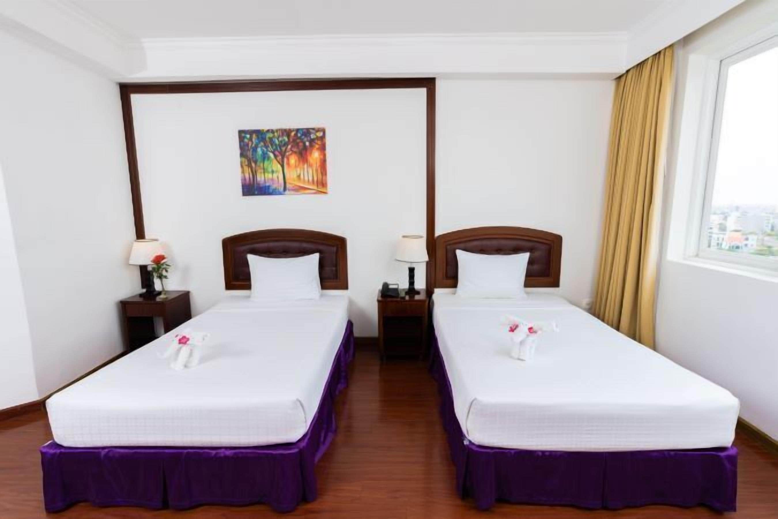 A hotel room with two single beds, a nightstand with a lamp, a painting on the wall, and a window with curtains.