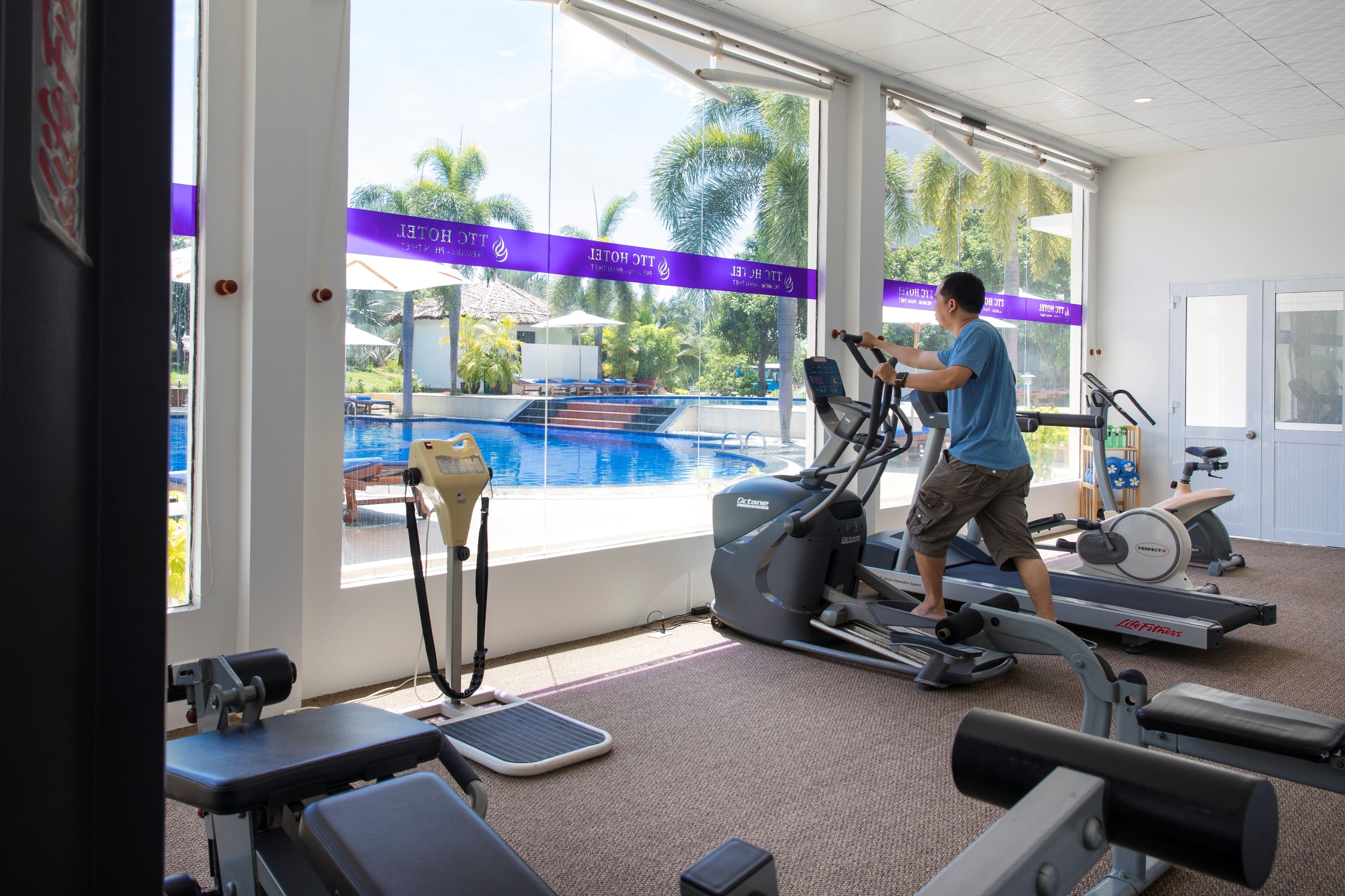 Un gimnasio con cintas de correr, una elíptica y una bicicleta estática. Un ventanal ofrece vistas a una piscina y palmeras.