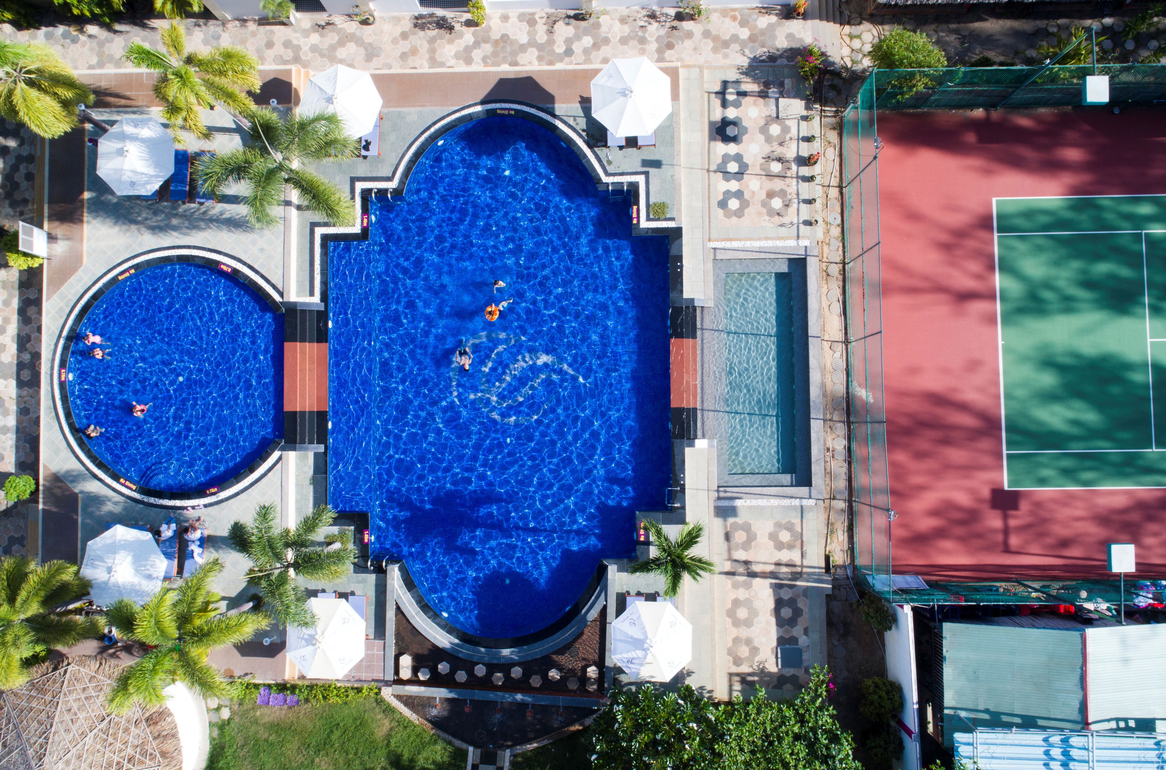 Vue aérienne d’un hôtel doté de deux piscines, d’un court de tennis et entouré de palmiers.