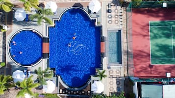 An aerial view of a hotel with two swimming pools, a tennis court, and surrounding palm trees.
