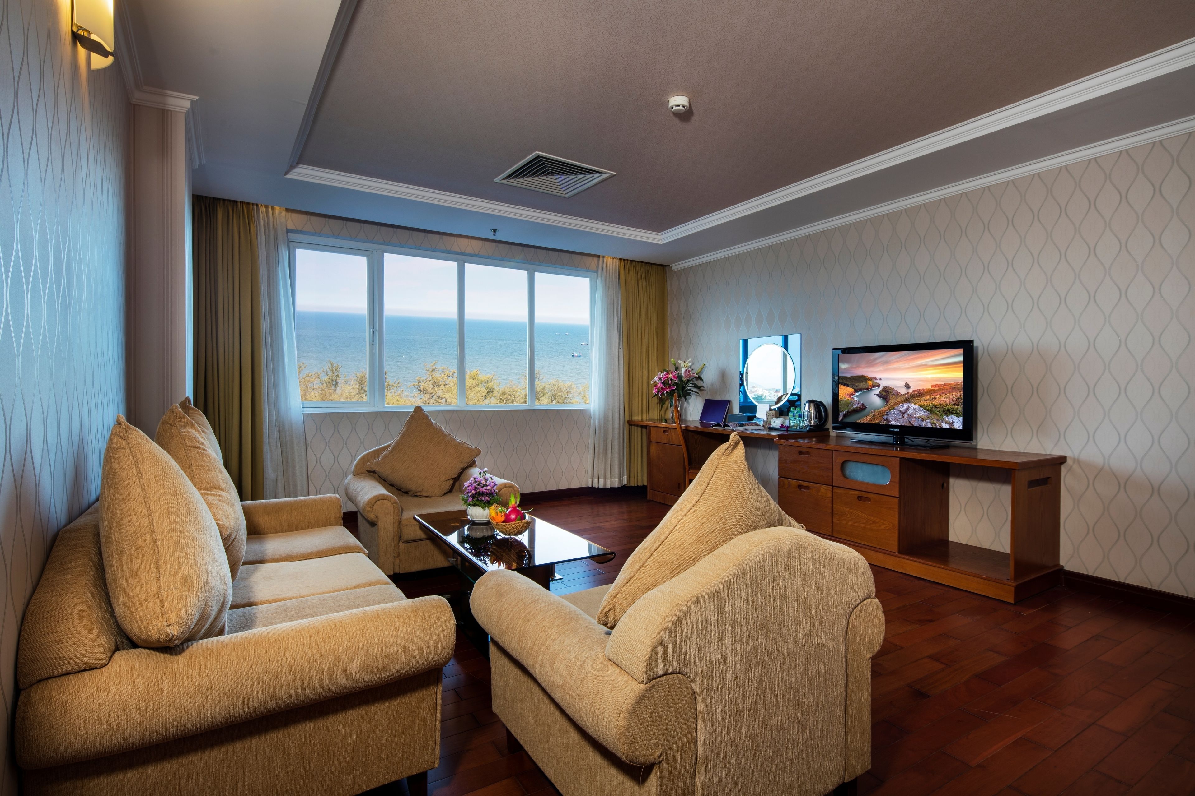 A hotel room with a sofa set, a television, a desk with a laptop, and a view of the sea.