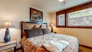 A bedroom with a large bed, two bedside tables with lamps, a ceiling fan, a stone wall, and a window with a view of greenery.