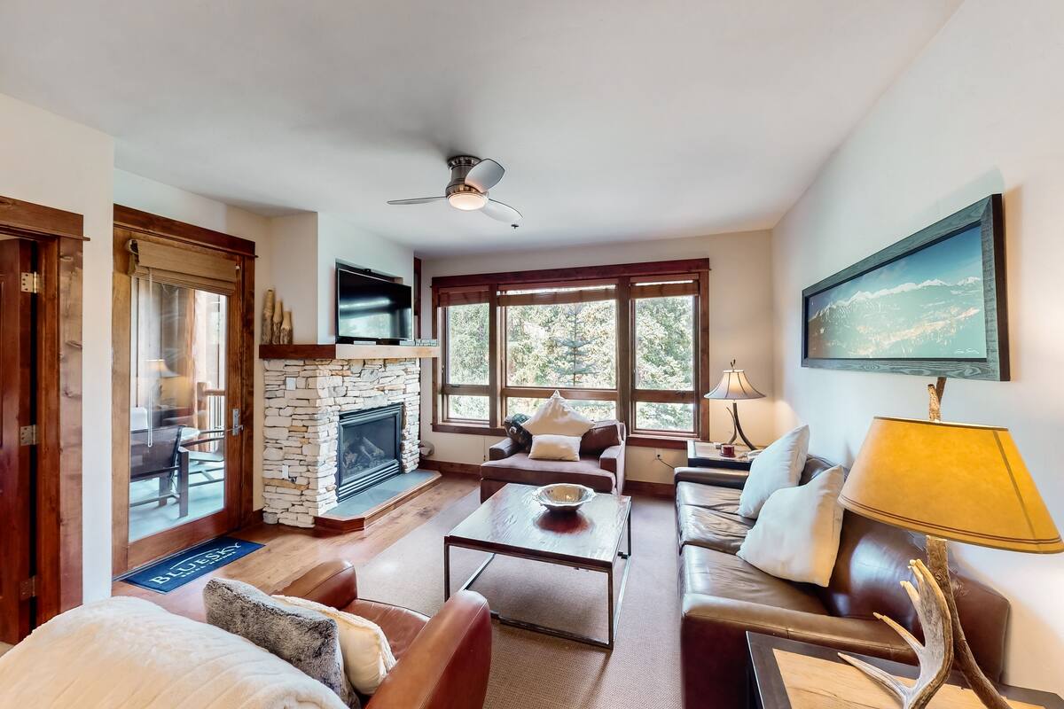 A cozy living room with a stone fireplace, leather sofas, a coffee table, and a large window with a view of trees.
