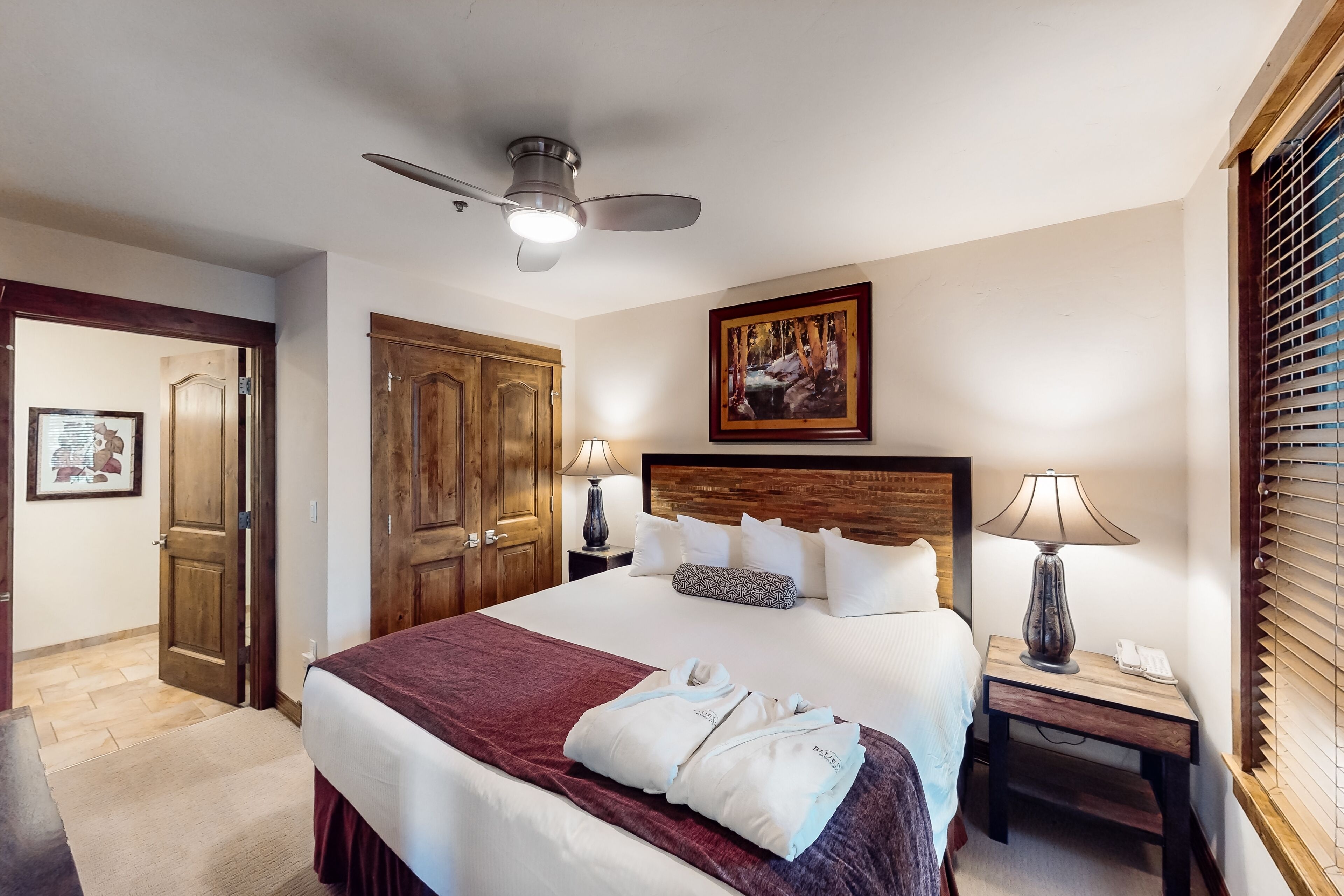 A hotel room with a large bed, wooden wardrobe, ceiling fan, bedside lamps, and a painting on the wall.