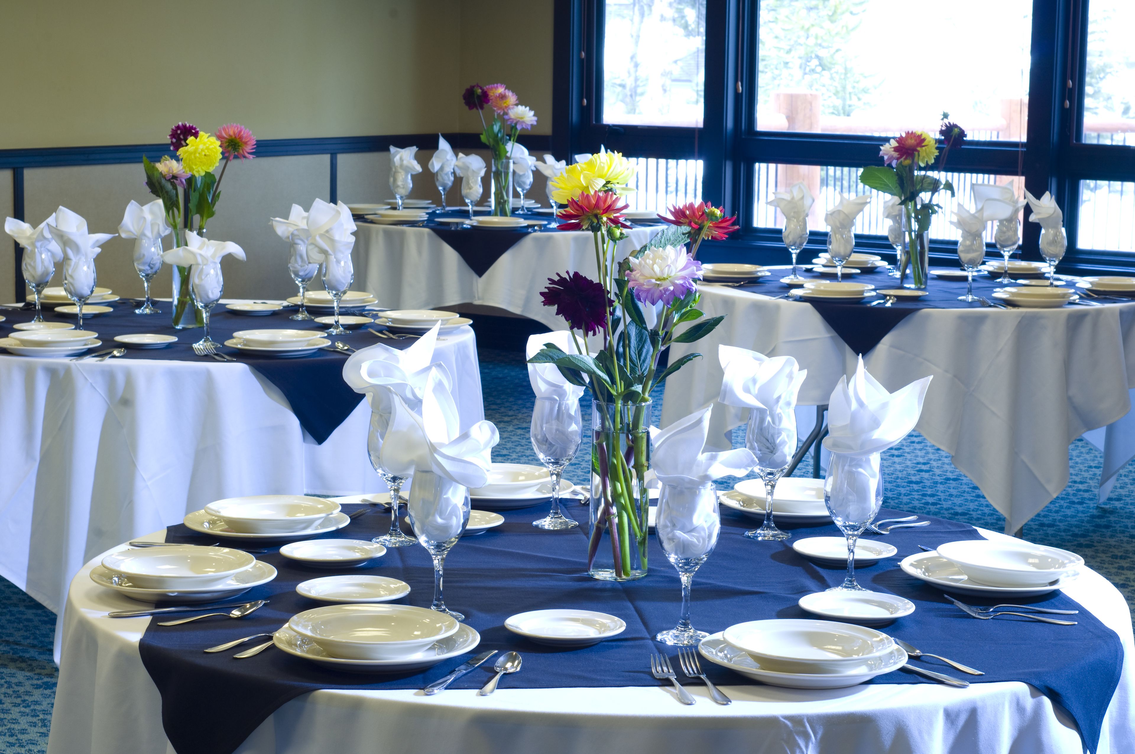A banquet hall with round tables set for dining, each with white plates, silverware, and glassware, and centerpieces of flowers in vases.