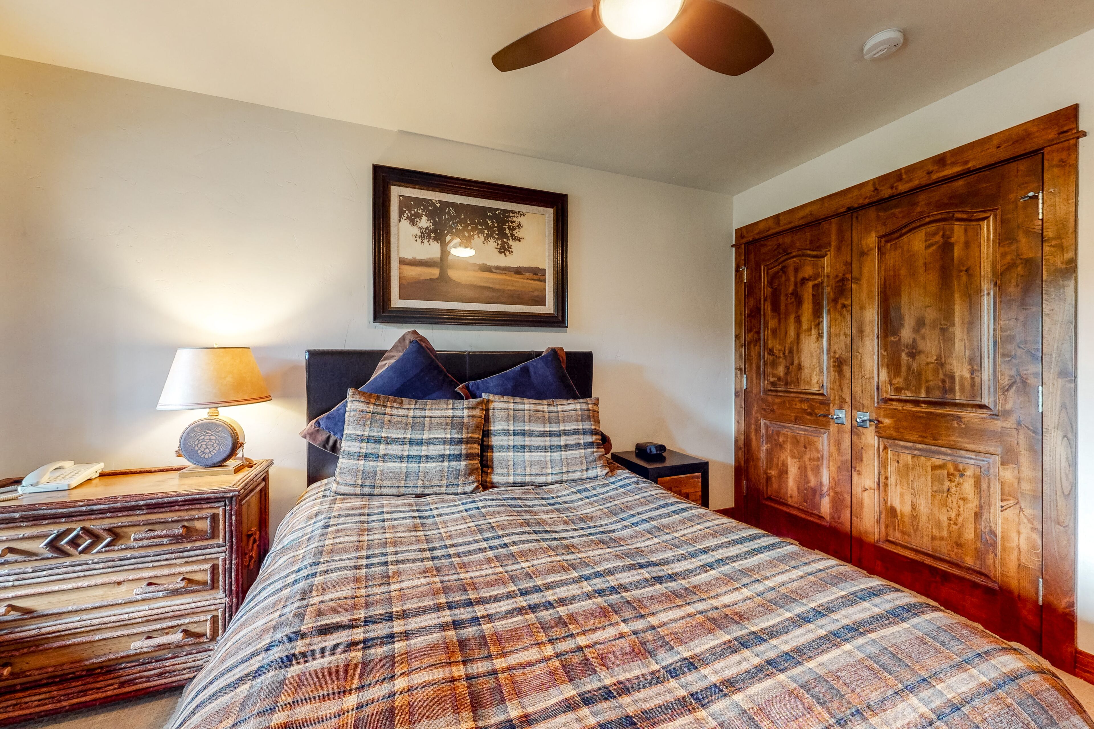 A bedroom with a plaid bedspread, a wooden headboard, a nightstand with a lamp, a framed picture on the wall, and a large wooden wardrobe.