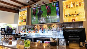 Uma área de bar com uma grande tela de televisão, prateleiras cheias de garrafas de bebidas e uma máquina de café.
