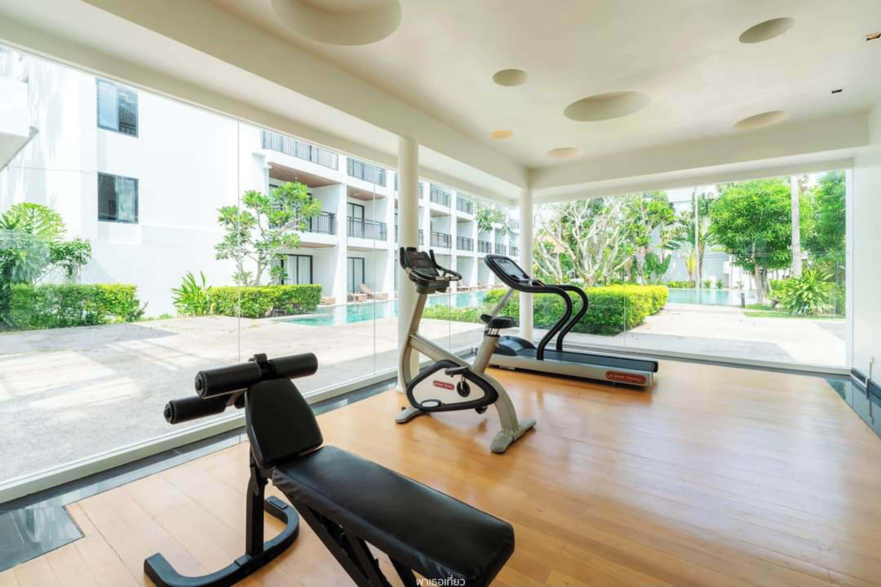 Un gimnasio con equipamiento de ejercicio, ventanas amplias y vistas a un edificio y vegetación.