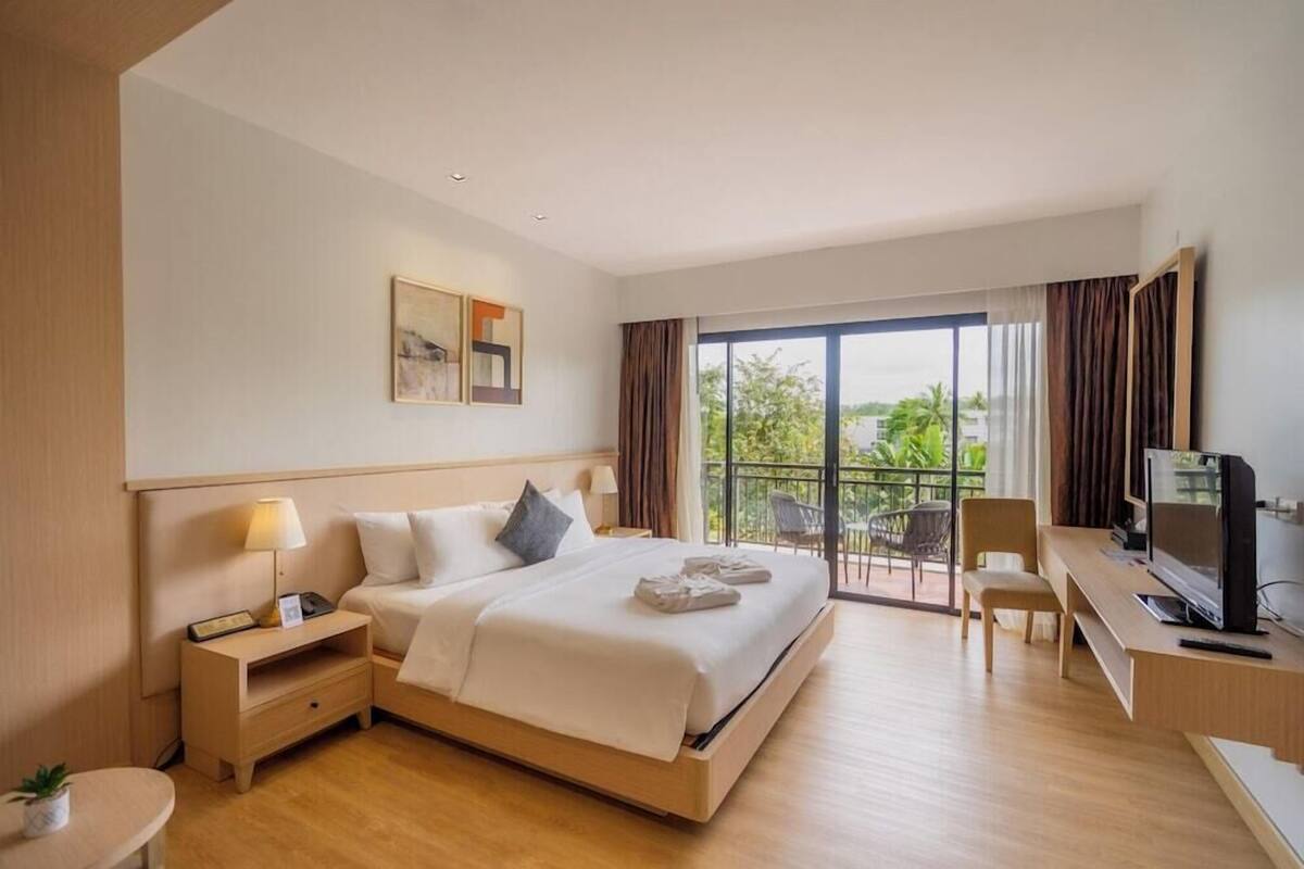 Habitación de hotel con cama, escritorio con televisor, silla, mesita de noche y balcón con vistas.