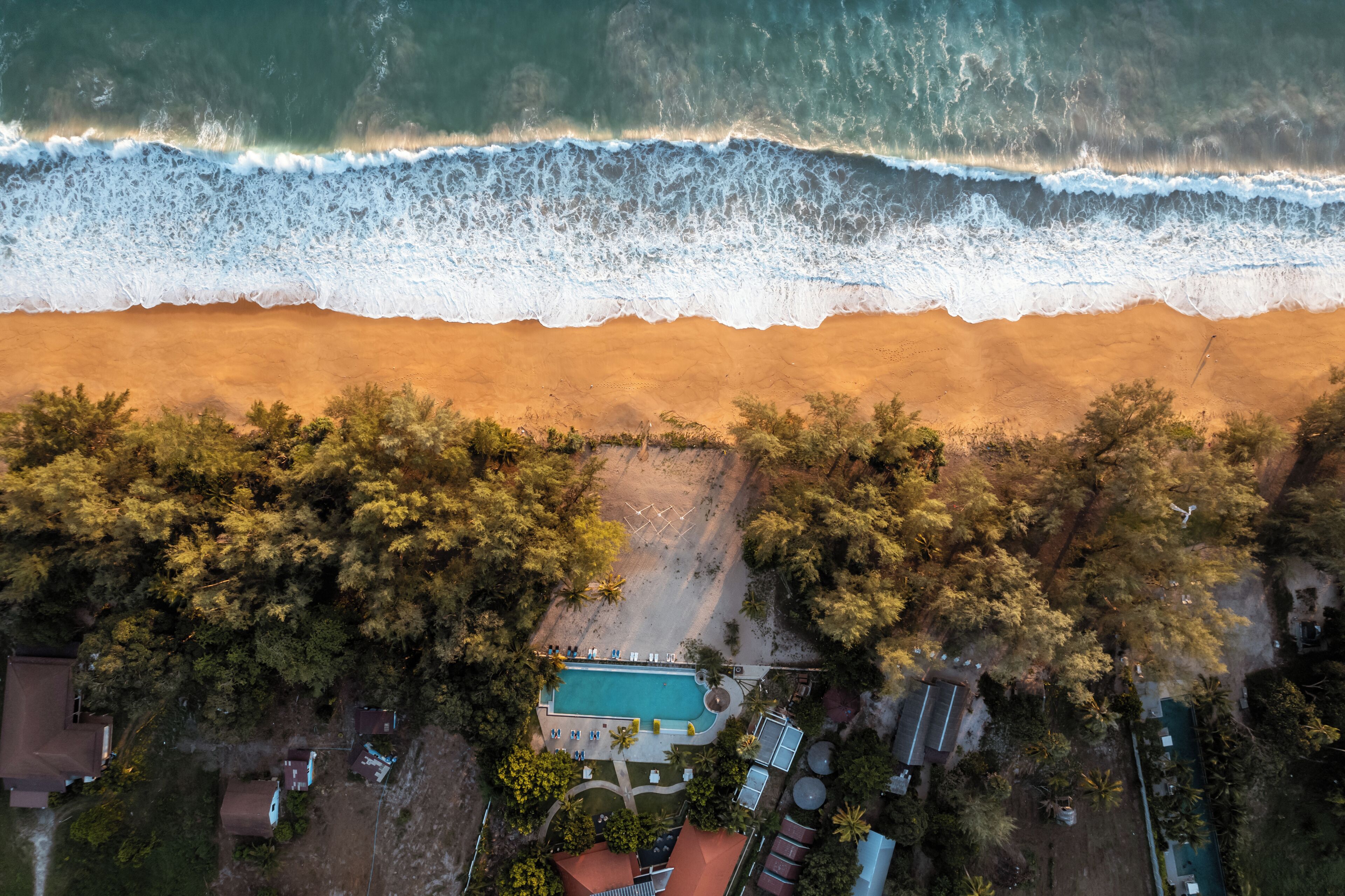 An aerial view of a beach with a swimming pool surrounded by trees and a few buildings.