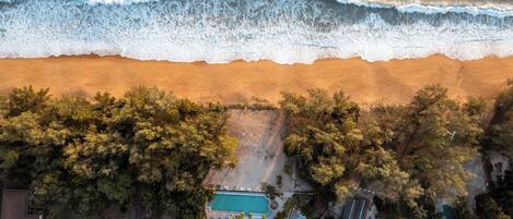 An aerial view of a beach with a swimming pool surrounded by trees and a few buildings.