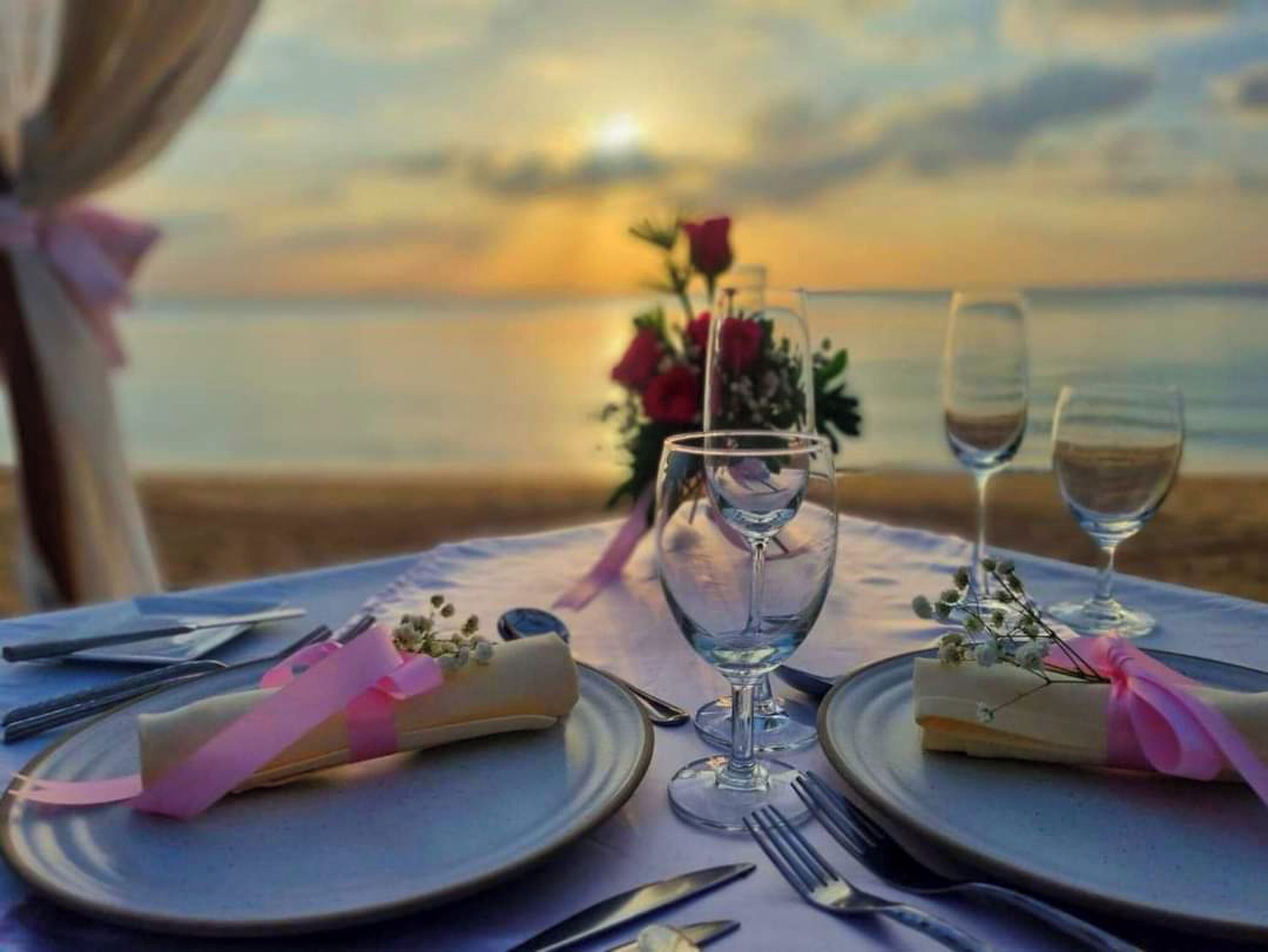 Une table dressée pour deux personnes, avec vue sur l’océan au coucher du soleil.