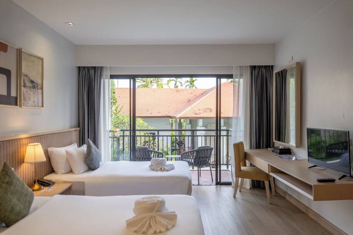 Habitación de hotel con dos camas, un escritorio, un televisor y un balcón con vistas a edificios y zonas verdes.
