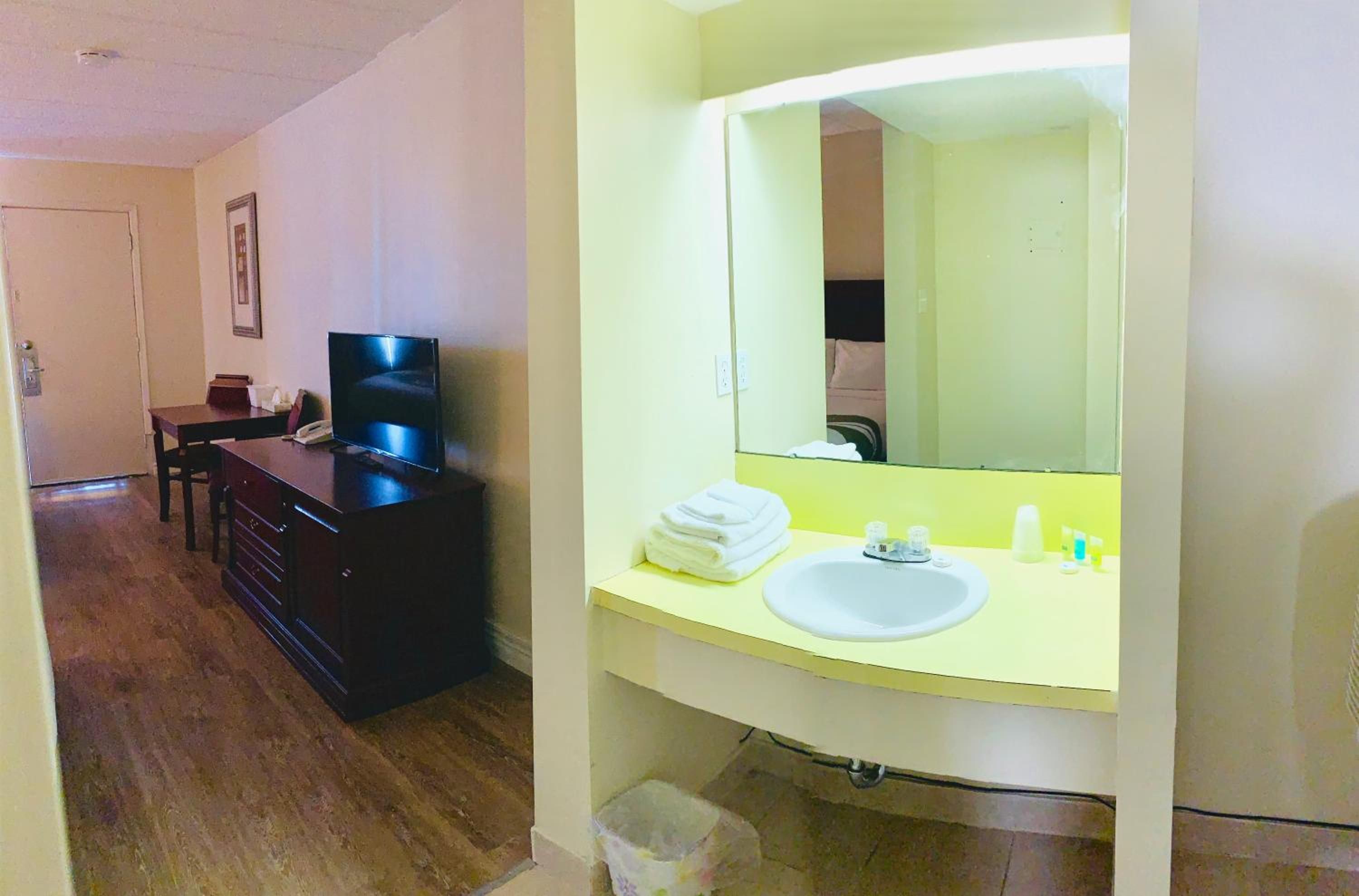 Habitación de hotel con piso de madera, cómoda de color oscuro, televisor, espejo grande y baño con lavamanos y artículos de aseo.