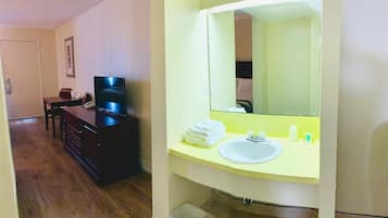 Habitación de hotel con piso de madera, cómoda de color oscuro, televisor, espejo grande y baño con lavamanos y artículos de aseo.