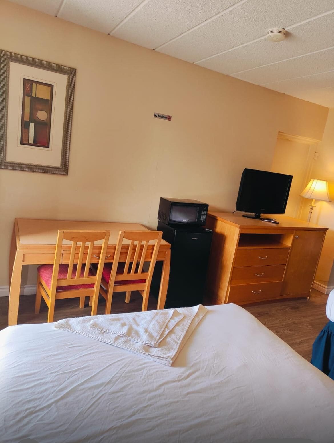 A hotel room with a bed, a desk with a television, a chair, a small refrigerator, and a 'No Smoking' sign on the wall.