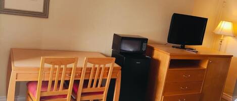 A hotel room with a bed, a desk with a television, a chair, a small refrigerator, and a 'No Smoking' sign on the wall.