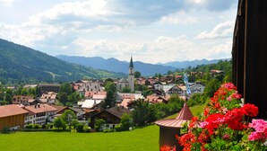 Une ville où se dresse une église imposante, entourée de maisons et de verdure, vue depuis un balcon orné de fleurs au premier plan.