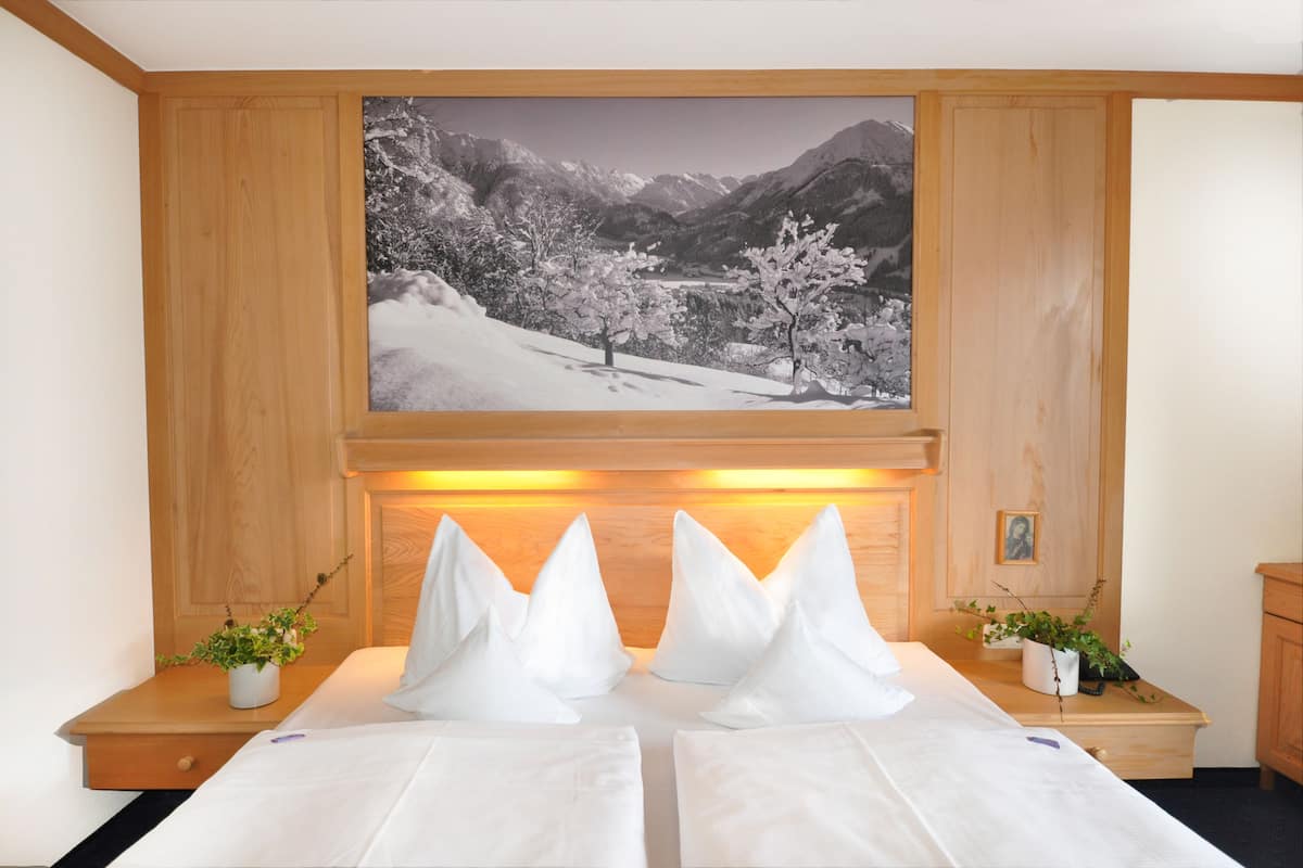 Een hotelkamer met een bed, twee nachtkastjes met planten en een groot schilderij van een besneeerd landschap, en houten lambrisering.