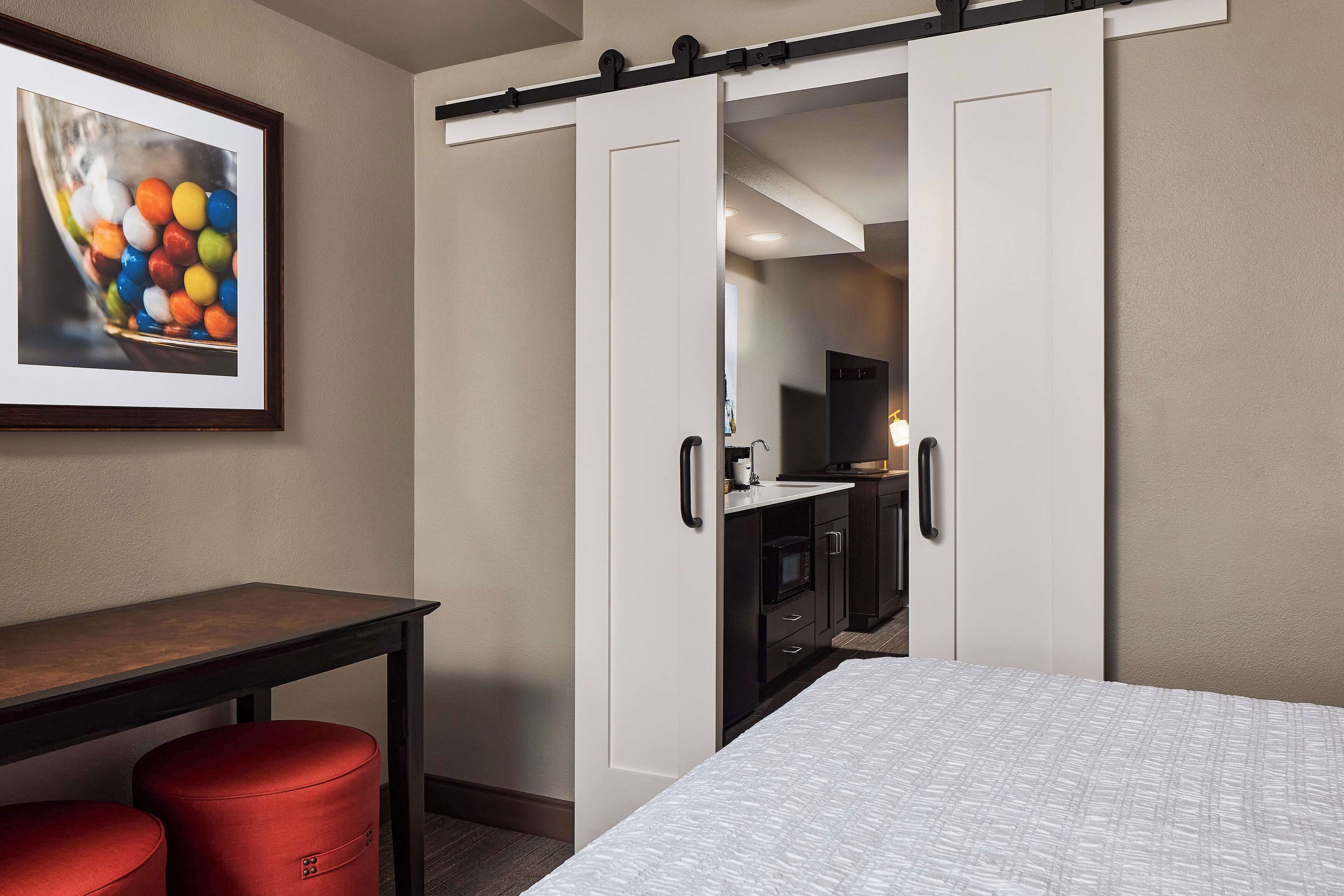 Ein Hotelzimmer mit Bett, Schreibtisch, einem roten Hocker, einem Kleiderschrank und einem gerahmten Bild an der Wand.