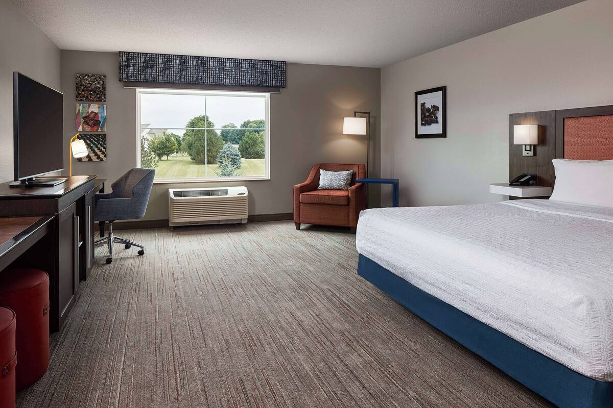 A hotel room with a bed, desk, chair, and a large window with a view of trees.