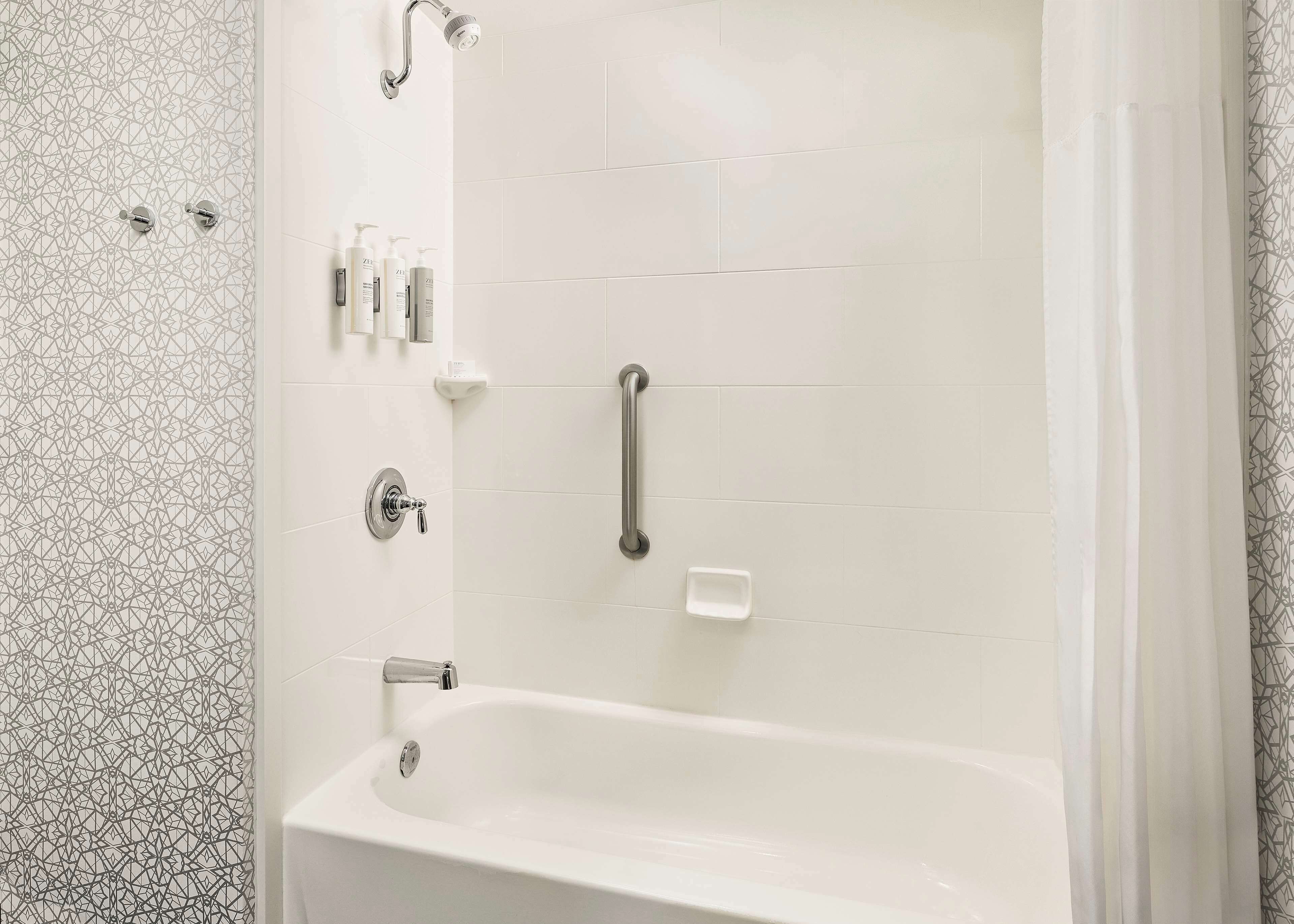 A bathroom with a white bathtub, a shower with a handheld showerhead, and a handrail. There are toiletries on the wall and a soap dish.