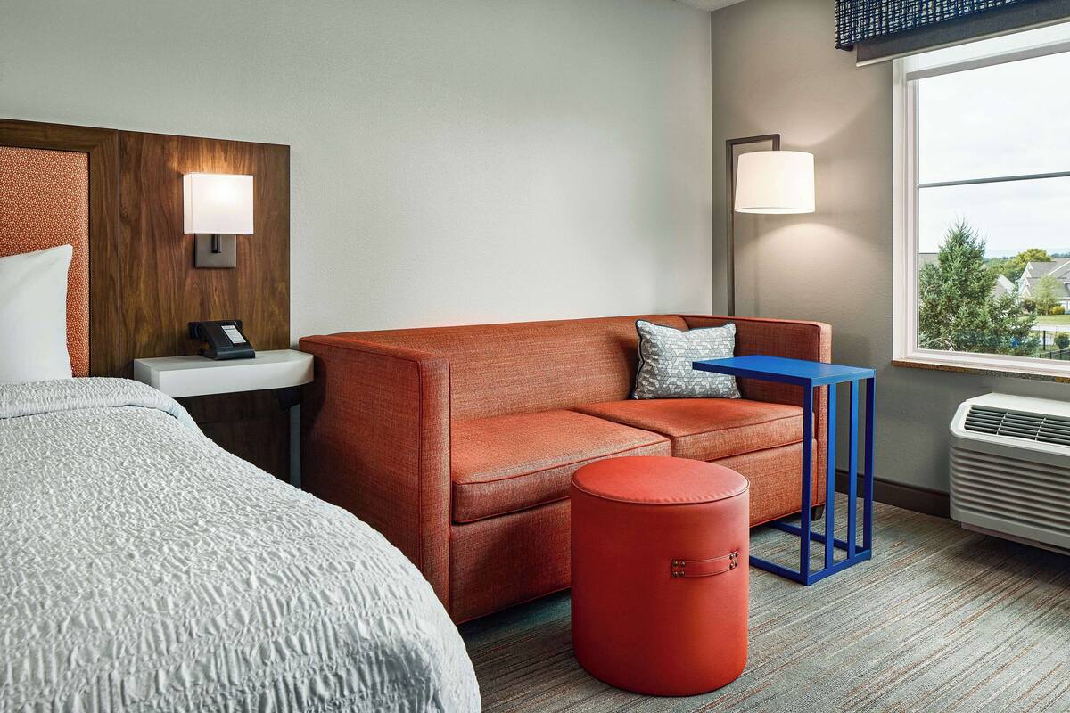 A hotel room with a bed, a sofa, a small table, a red ottoman, a wall-mounted lamp, and a window with a view.