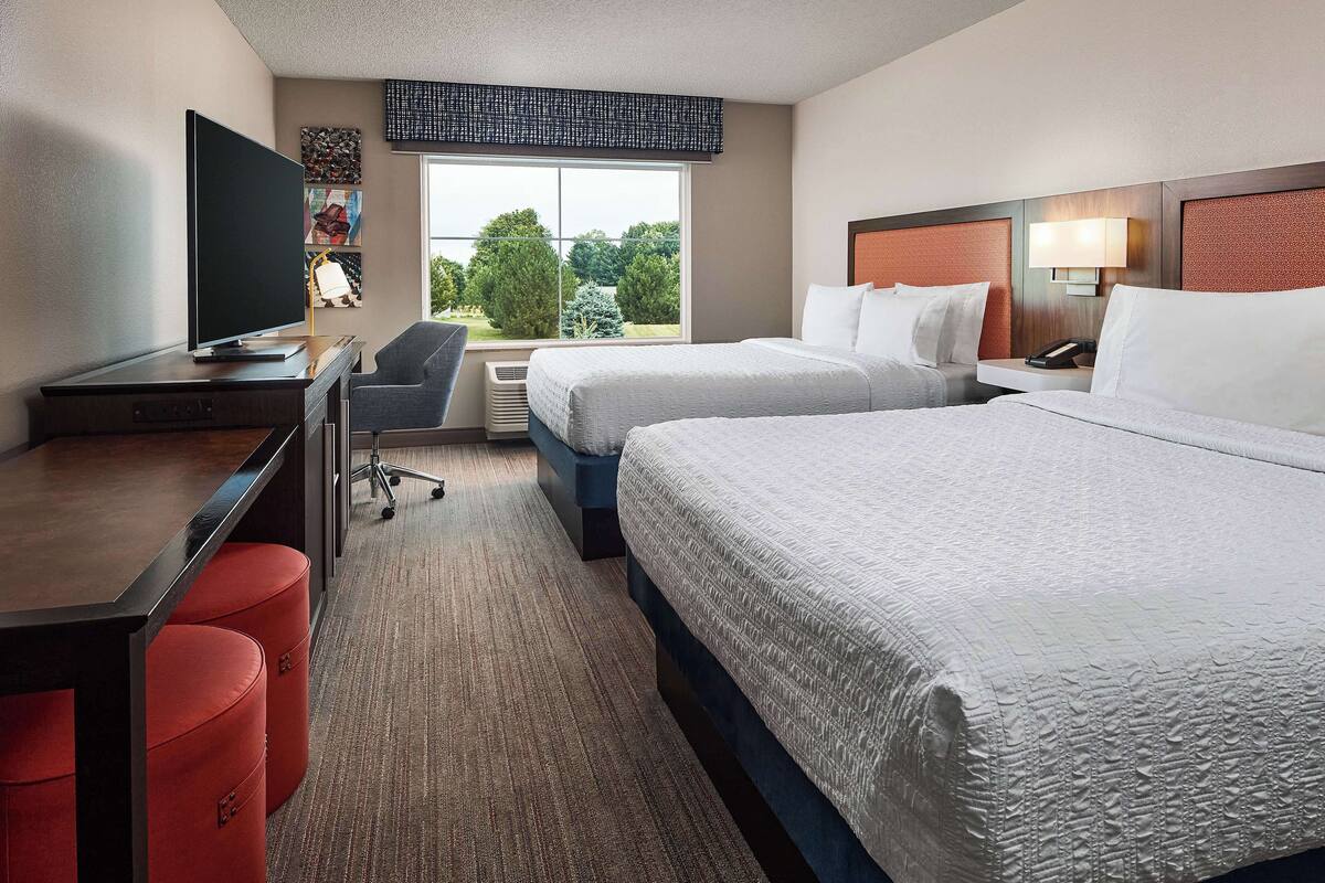 A hotel room with two beds, a desk, a chair, a TV, and a window with a view of trees.