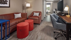 A hotel room with a red sofa, a blue armchair, a grey office chair, a flat-screen TV, a lamp, and a framed picture on the wall.