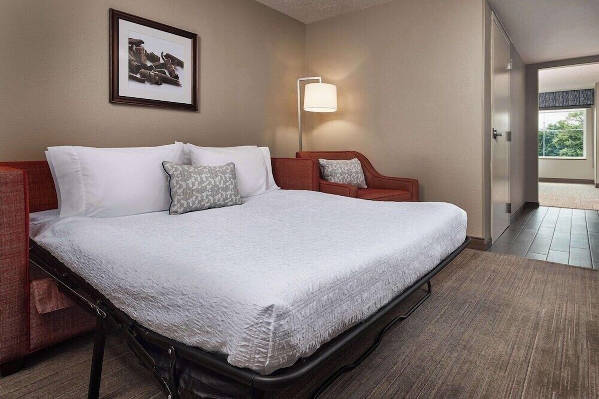 A hotel room with a large bed, a red armchair, a standing lamp, and a framed picture on the wall.