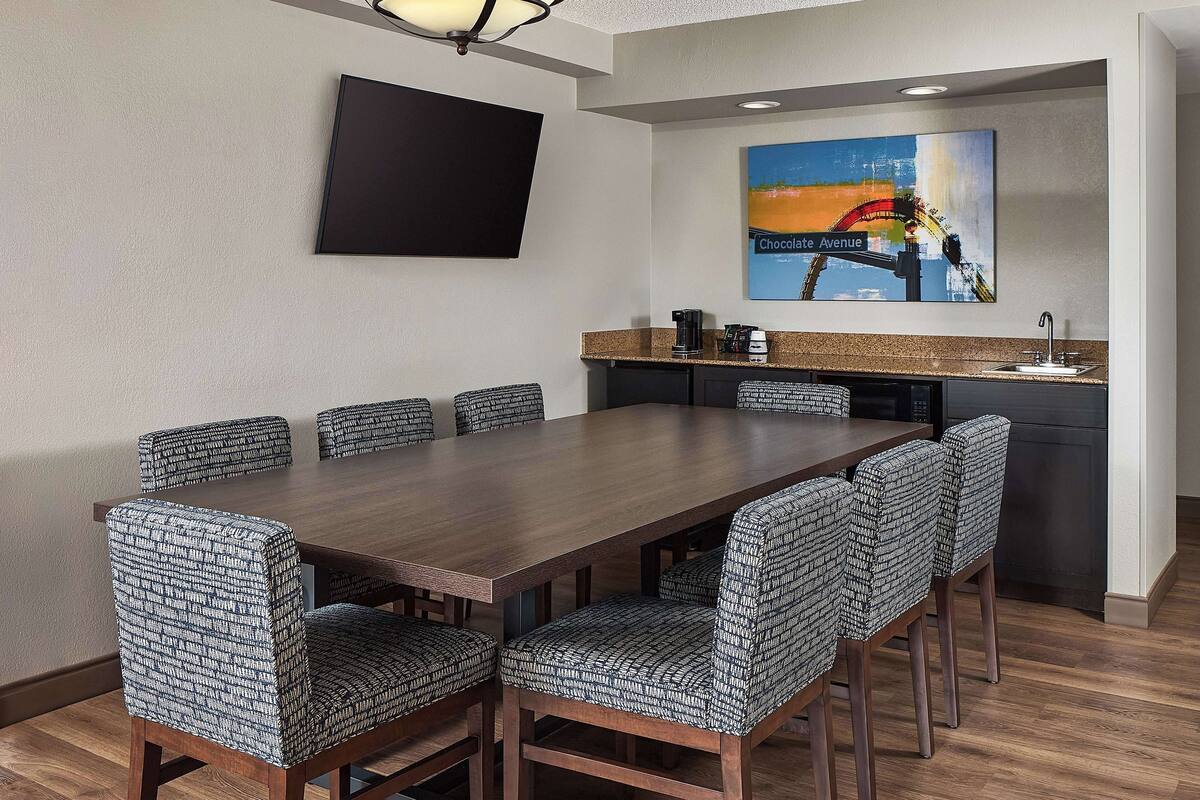 A conference room with a long wooden table and patterned chairs, a kitchen area with a sink and coffee maker, and a painting of a bridge on the wall.
