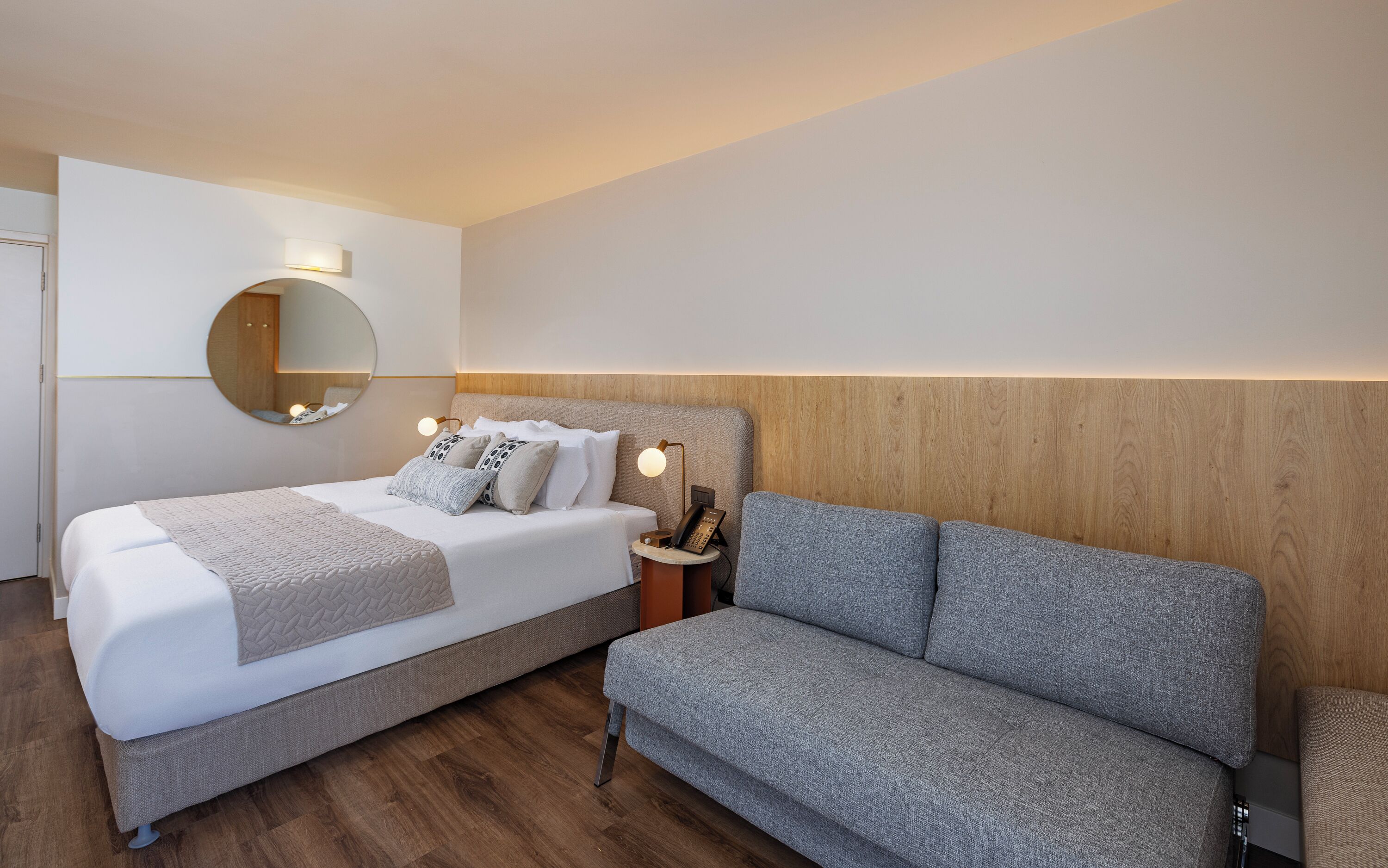 A modern hotel room with a bed, a sofa, a bedside table with a lamp, and a round mirror on the wall.