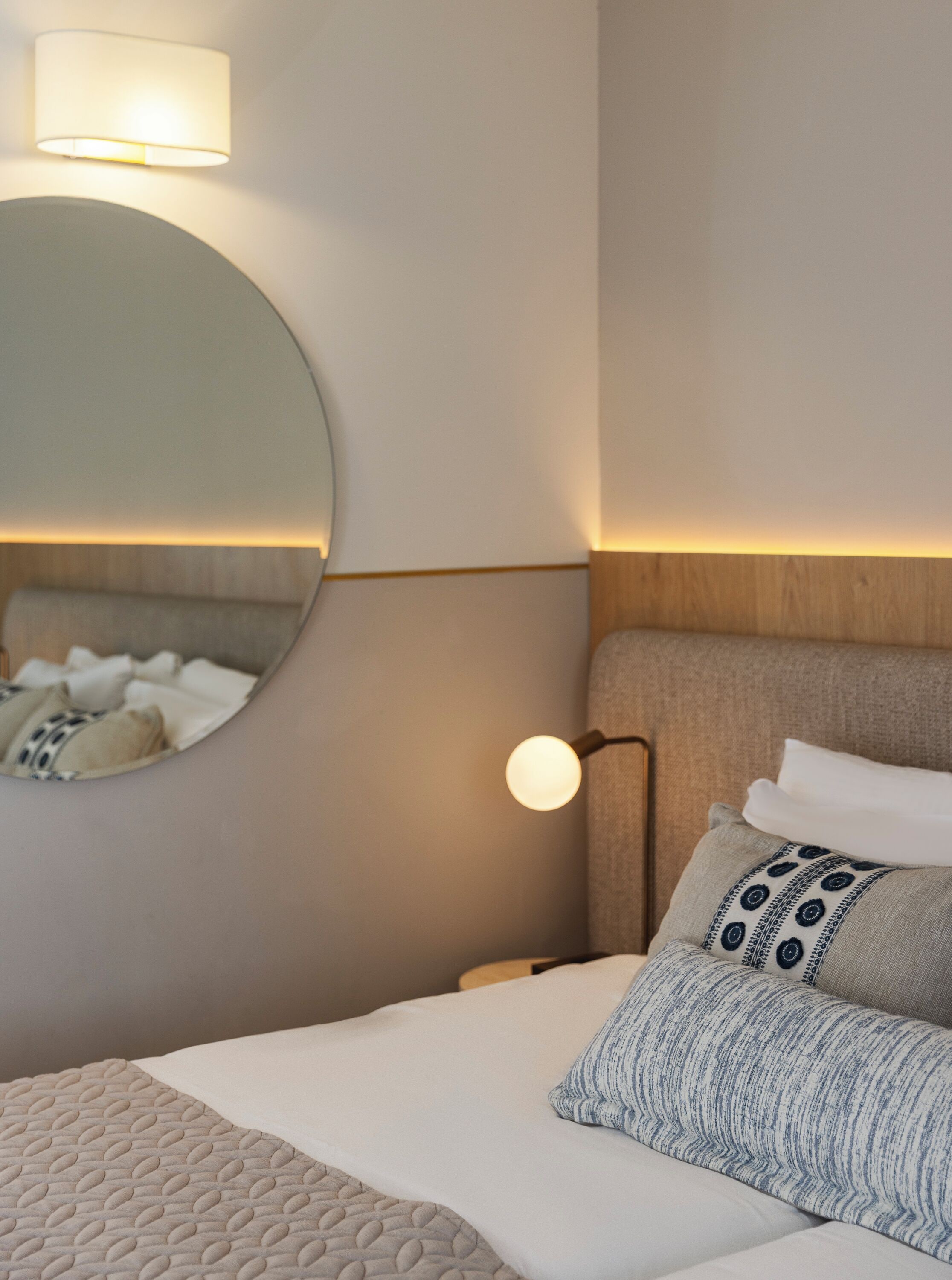 En pænt redt seng med et vamset tæppe, et rundt spejl og en lampe på væggen.