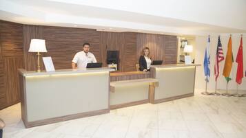 A reception desk with two staff members, a wooden wall, and flags.