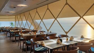 A restaurant with tables set for dining, chairs, and a view of the sea.