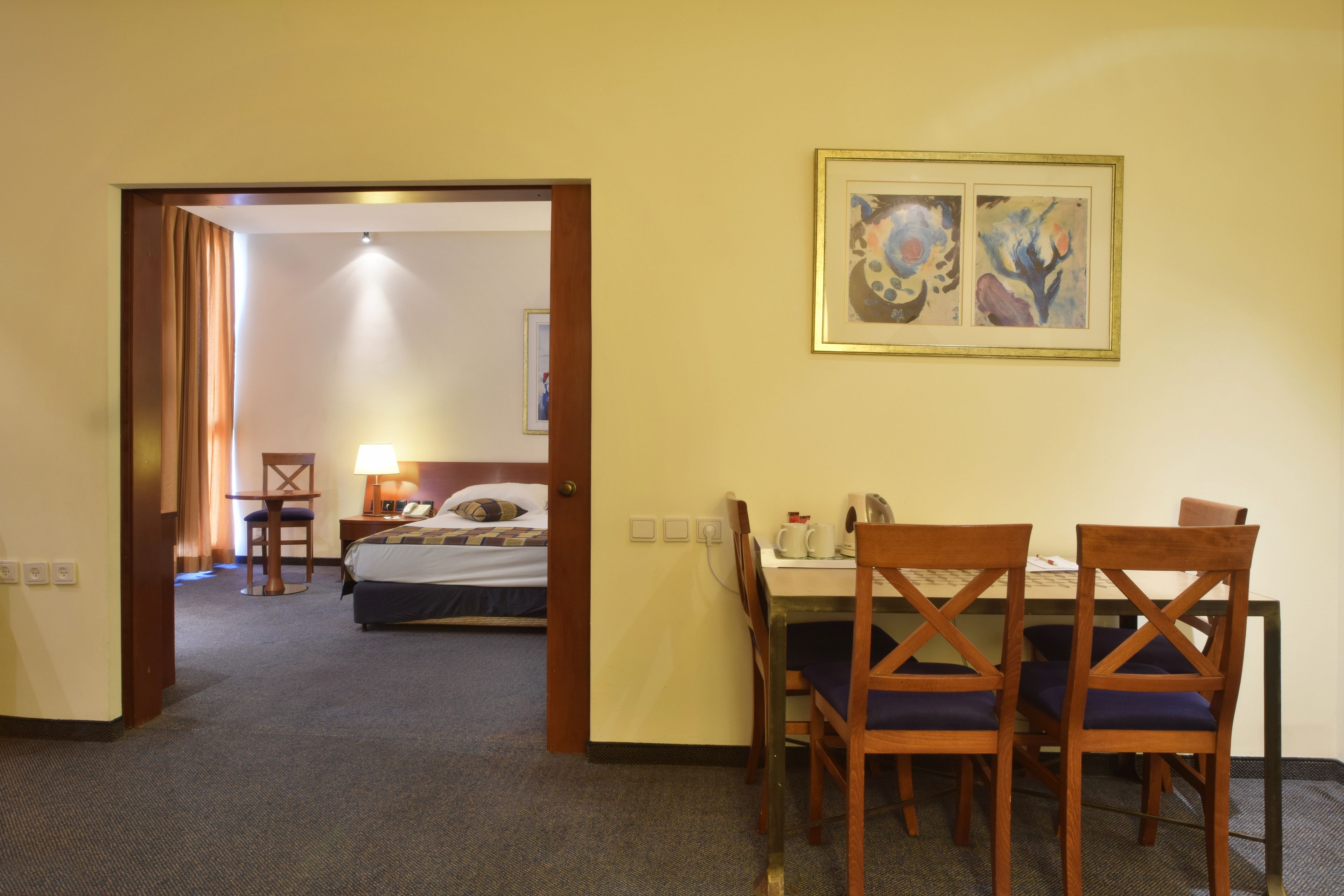 Une chambre d’hôtel avec un lit, un bureau avec des chaises, un tableau au mur et une porte donnant sur une autre pièce.