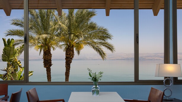 Una habitación con una mesa y sillas, un jarrón con flores, y vistas a palmeras y una masa de agua a través de amplias ventanas.